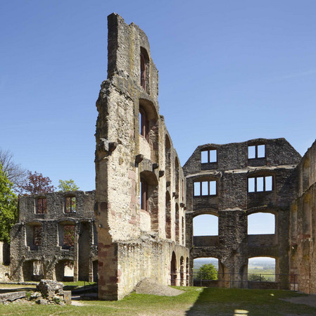 Mauerwände mit Fensteröffnungen, links vorne ein Brunnen, im Hintergrund Bäume